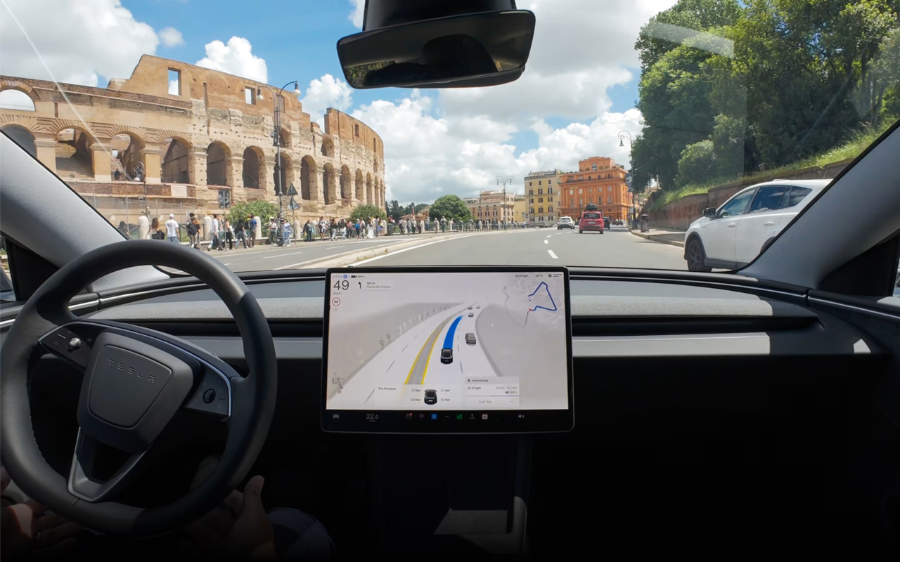 Image - Not an Fsd Tracker Inside a Tesla, Dashboard Centered on a Navigation Map While the Road and a Historic Stone Amphitheater Line the Street on the Left, with Pedestrians and Colorful Buildings Ahead Under a Blue Sky.